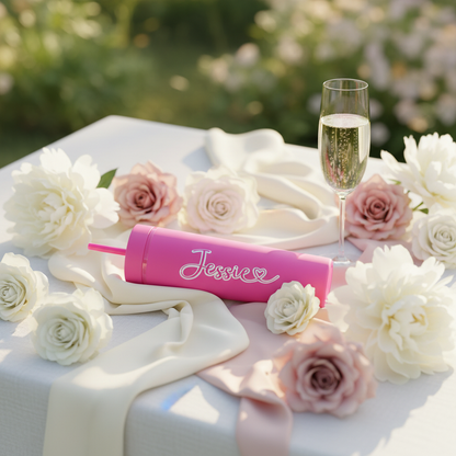 Personalised hot pink cold cup with custom name, handmade in UK, on white tablecloth surrounded by blush roses, white peonies, and champagne flute in outdoor garden setting