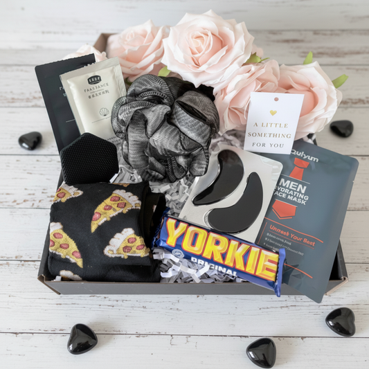 Open black pamper gift box for him containing pizza socks, Yorkie chocolate bar, grey bath puff, eye gel pads, men's hydrating face mask and travel sachets pamper gift UK