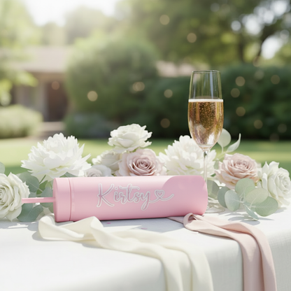 Personalised pink cold cup with custom name, handmade in UK, on white tablecloth surrounded by blush roses, white peonies, and champagne flute in outdoor garden setting